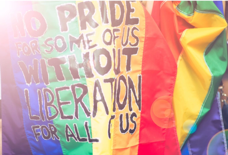 Colorful pride flag behind a sign calling for liberation for all LGBTQ+ individuals.