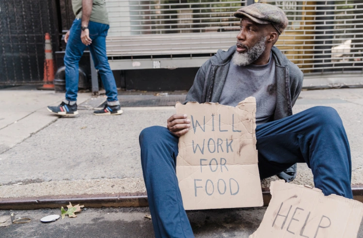Man sitting on sidewalk holding a sign that says 'Will work for food'.