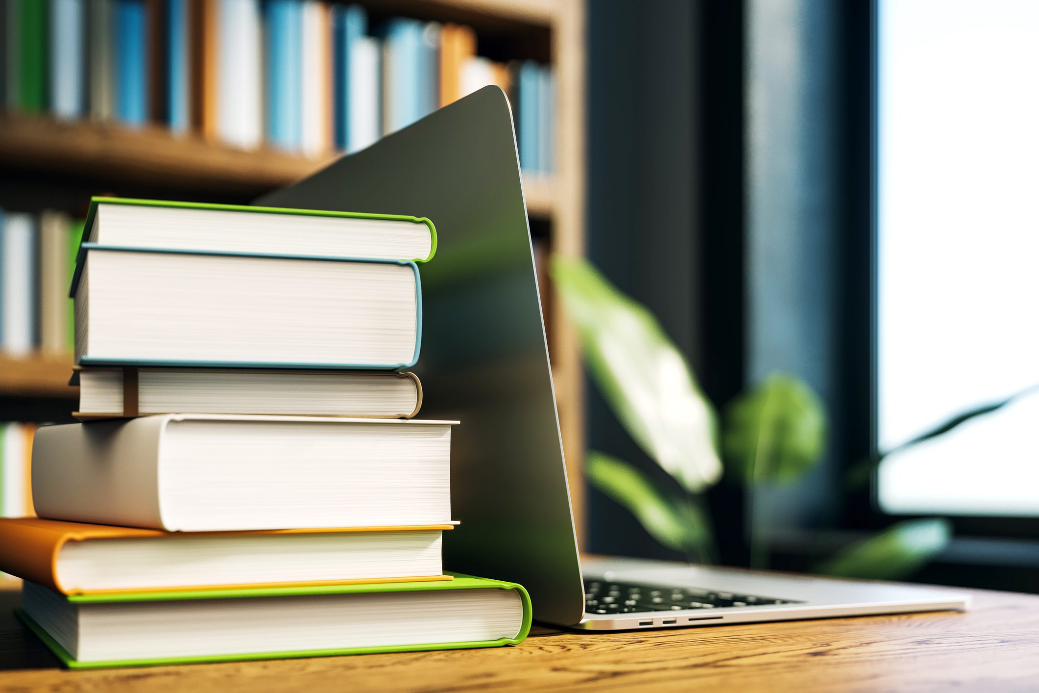 Stack of books leaning against a laptop on a wooden desk near a window.